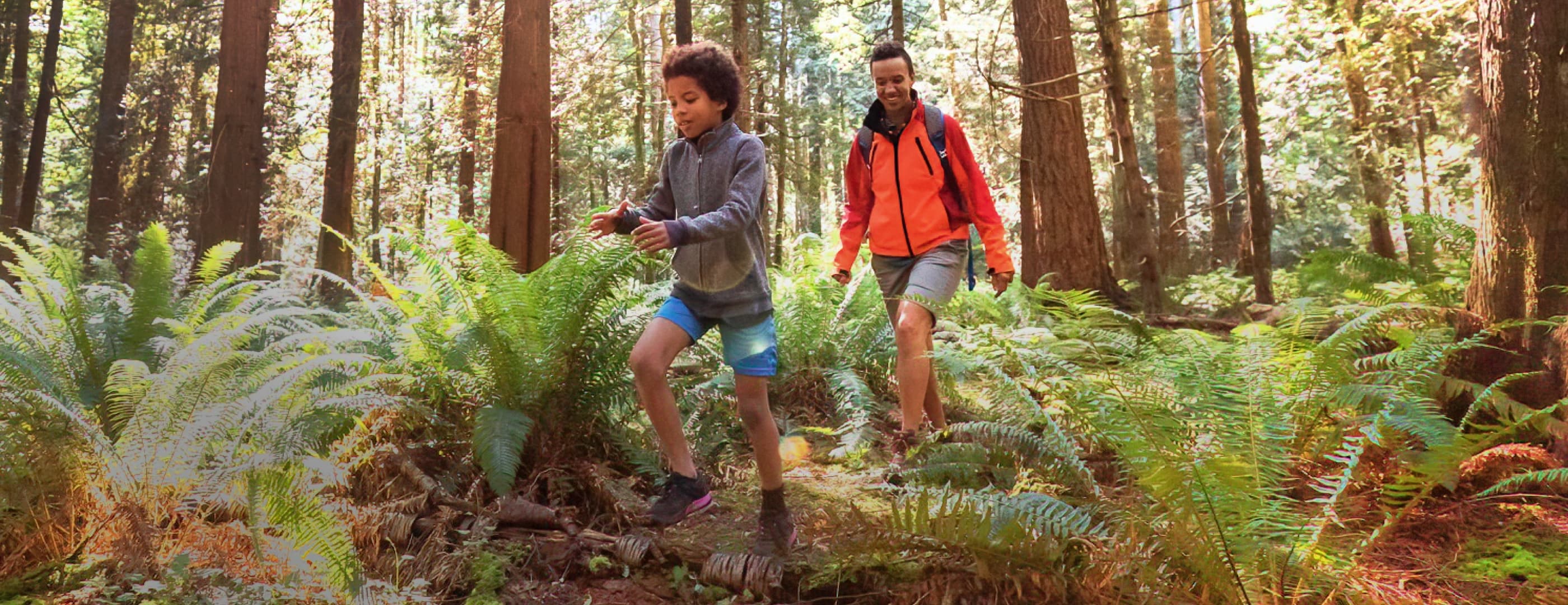 An adult in an orange raincoat and a child in a grey sweatshirt with dark hair are hiking through a forest, the adult smiles as the child skips over a small log on the trail.