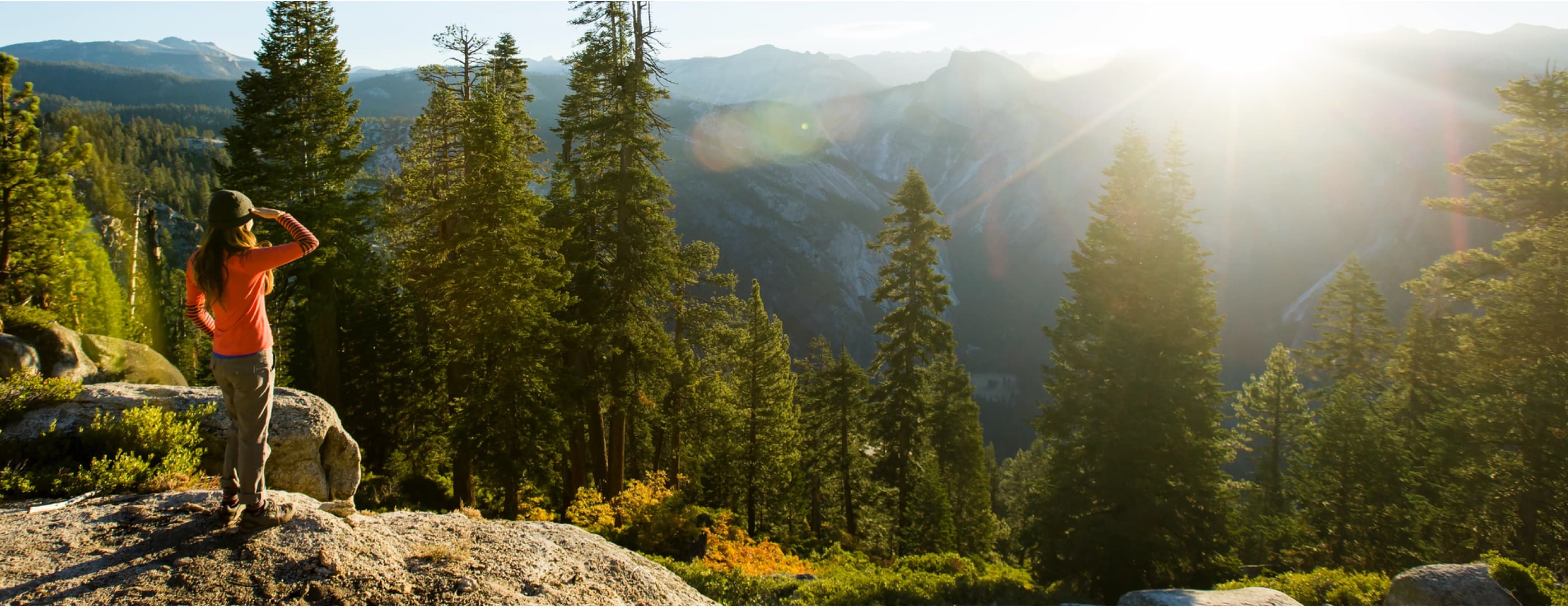 A hiker with long brown hair under a green beanie and an orange shirt looks over a tree-filled valley, shielding their eyes with their hand from the sun.