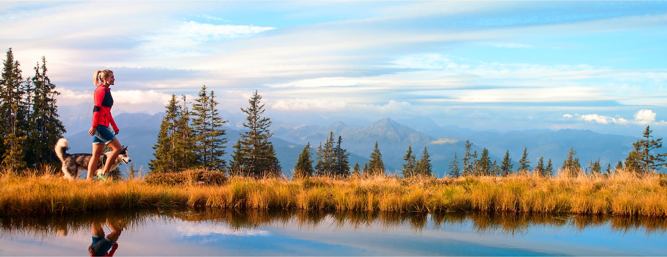 A hiker with a blond ponytail walks in front of a white husky dog through dry yellow grass and scattered trees alongside calm waters showing their reflection below.