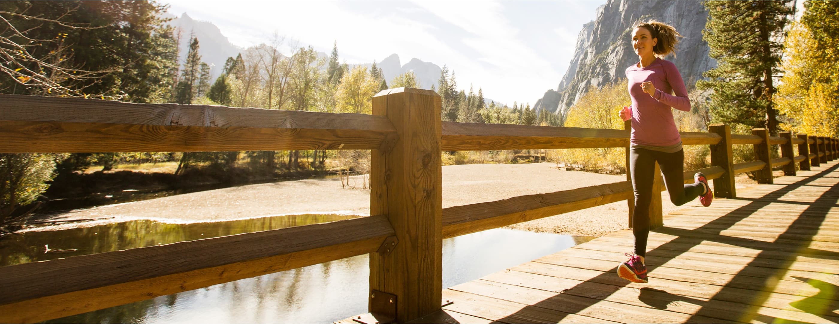 A runner with curly brown hair in a ponytail wearing a pink sweatshirt crosses a boardwalk bridge in front of towering rocky peaks.